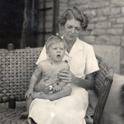 A black-and-white photograph of Mary Koch in a white dress sitting on a wicker chair holding a young Charles Koch in her lap.
