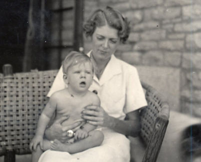 A black-and-white photograph of Mary Koch in a white dress sitting on a wicker chair holding a young Charles Koch in her lap.