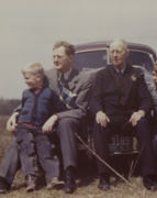 A colorized photo of a young Charles Koch standing in front of his father Fred Koch, seated alongside grandfather Harry Koch.