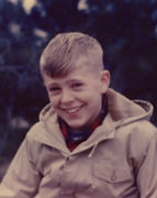 A vintage photo of a smiling young Charles Koch with light-colored hair wearing a tan hooded jacket over a red flannel shirt.