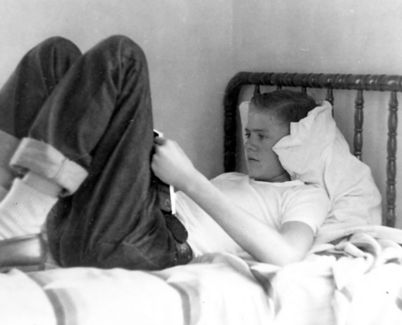 A black-and-white photo of an adolescent Charles Koch reading in bed, wearing rolled-up jeans and a white t-shirt.