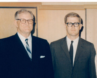 A vintage color photograph of Charles and Fred Koch standing side by side wearing horn-rimmed glasses and formal attire.