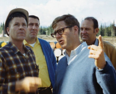 A vintage color photo of Charles Koch wearing glasses and a blue sweater speaking with three other men in an outdoor setting.