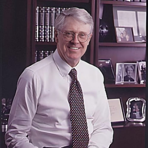 A photograph of Charles Koch in his office wearing a white shirt and patterned tie, seated in front of a bookshelf.