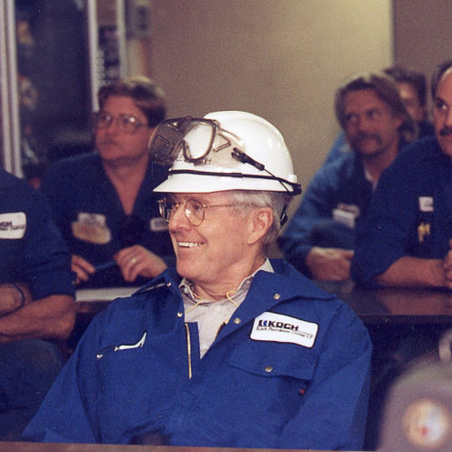 An image of Charles Koch wearing blue Koch-branded coveralls and a white hard hat with safety goggles resting on top.