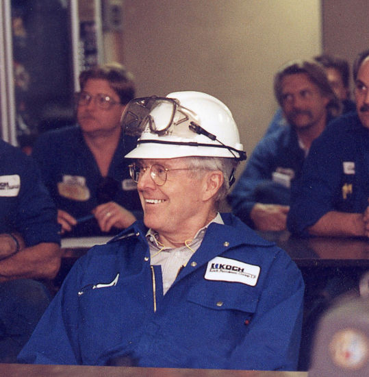 An image of Charles Koch wearing blue Koch-branded coveralls and a white hard hat with safety goggles resting on top.