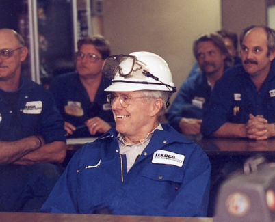 An image of Charles Koch wearing blue Koch-branded coveralls and a white hard hat with safety goggles resting on top.