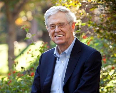 A photo of Charles Koch with white hair and glasses, wearing a navy blue blazer and light blue dress shirt, sitting outdoors.