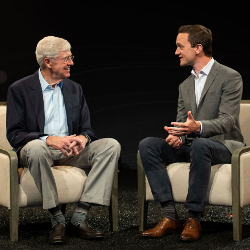 A photo of Charles Koch and Brian Hooks dressed in casual business attire seated on a stage discussing their book.
