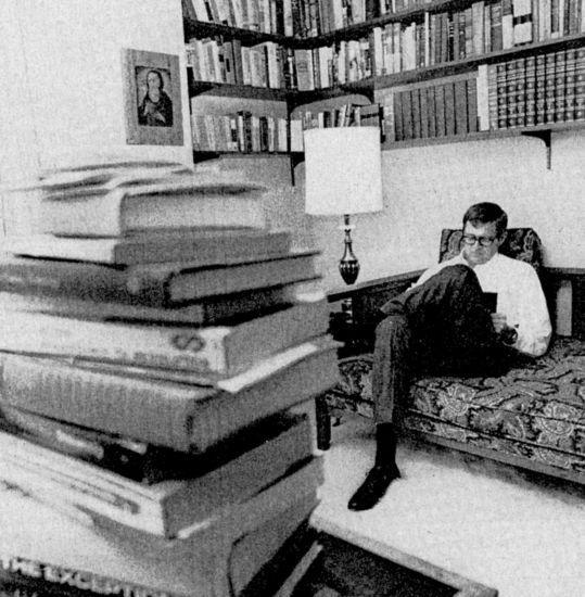 : A vintage black-and-white image of Charles Koch working in his study with a stack of books in the foreground.