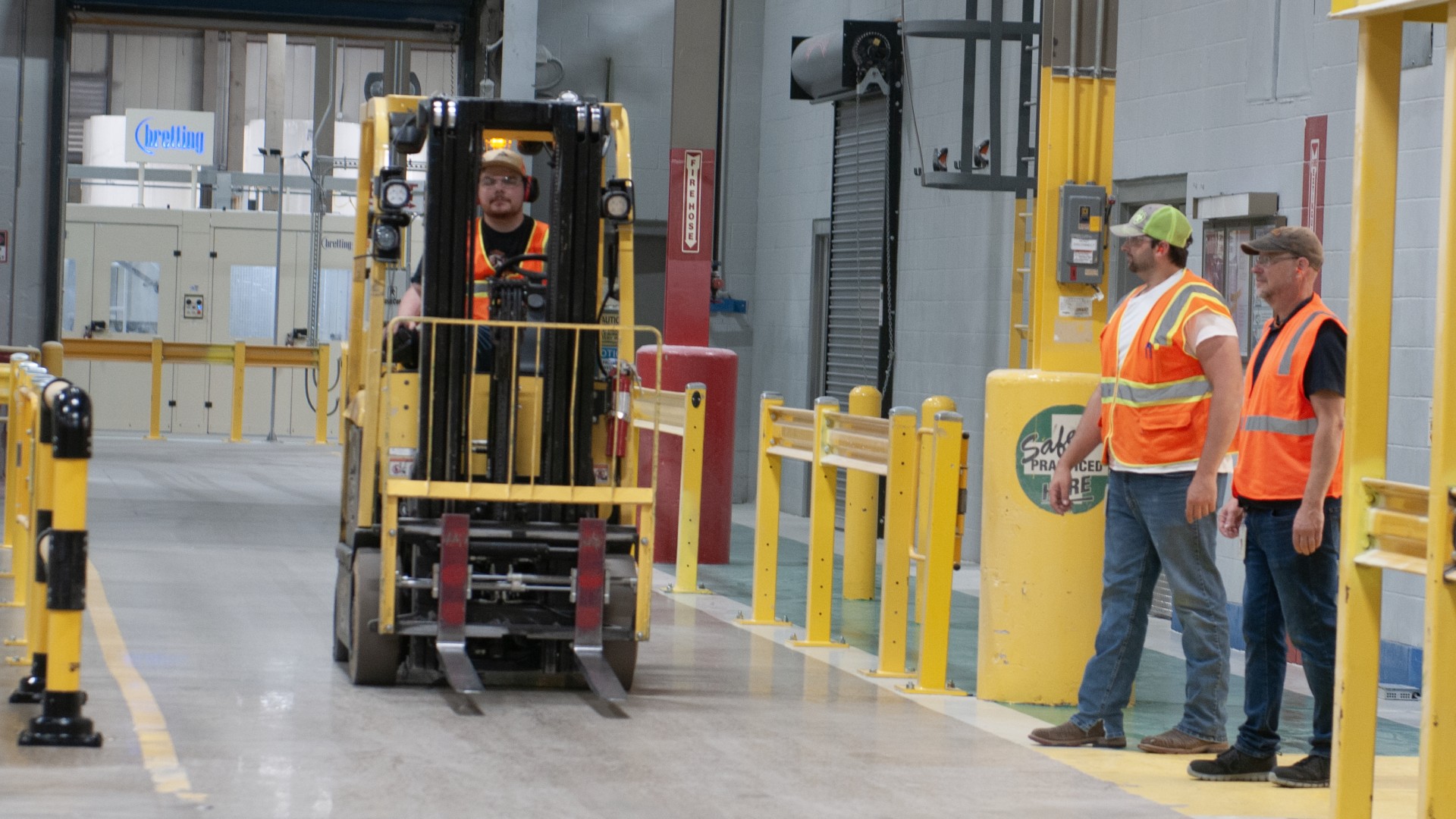 Employees stand near a fork lift lane wearing orange hi-vis safety equipment.