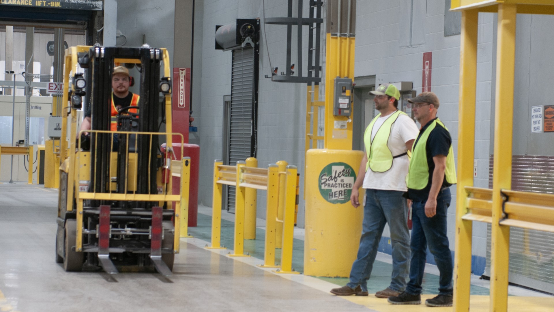 Employees stand near a fork lift lane wearing yellow hi-vis safety equipment.